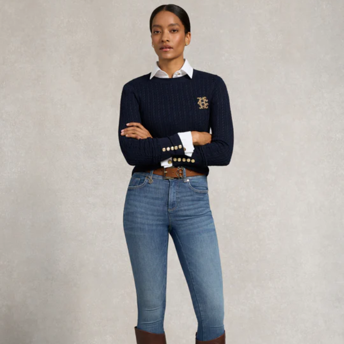 Woman wears blue skinny jeans with white shirt and a dark knitted top on a grey background
