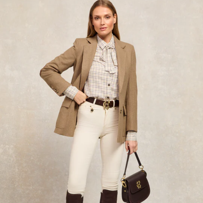 Woman wearing a brown blazer, white shirt, and white jeans, holding a leather handbag on a beige background