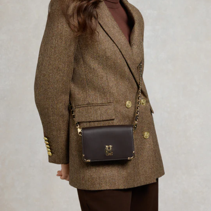 Person wearing a brown blazer with gold buttons and carrying a matching brown handbag on a plain background