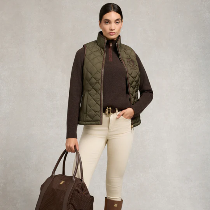 Woman wearing a green quilted gilet over a brown sweater, holding a brown bag on a light grey background