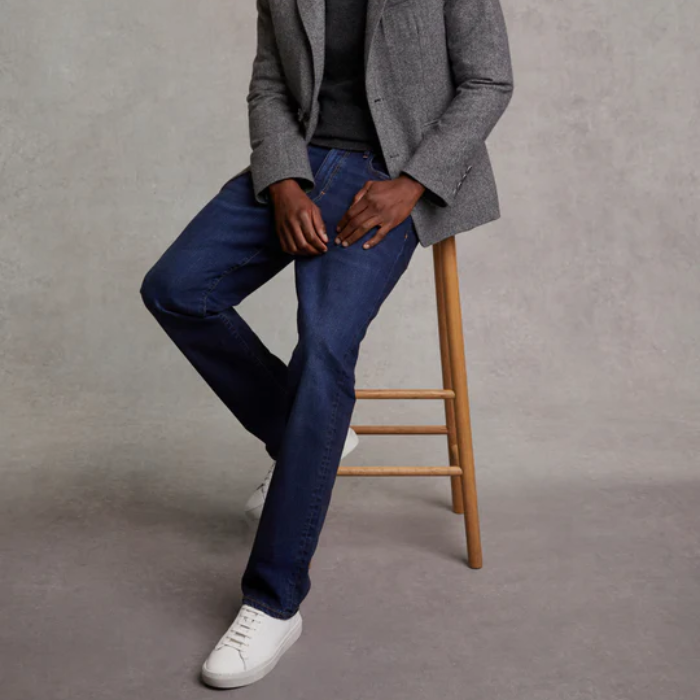 Person wearing a grey blazer, black shirt, blue jeans, and white training shoes on a wooden stool against a grey background.