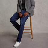 Person wearing a grey blazer, black shirt, blue jeans, and white training shoes on a wooden stool against a grey background.