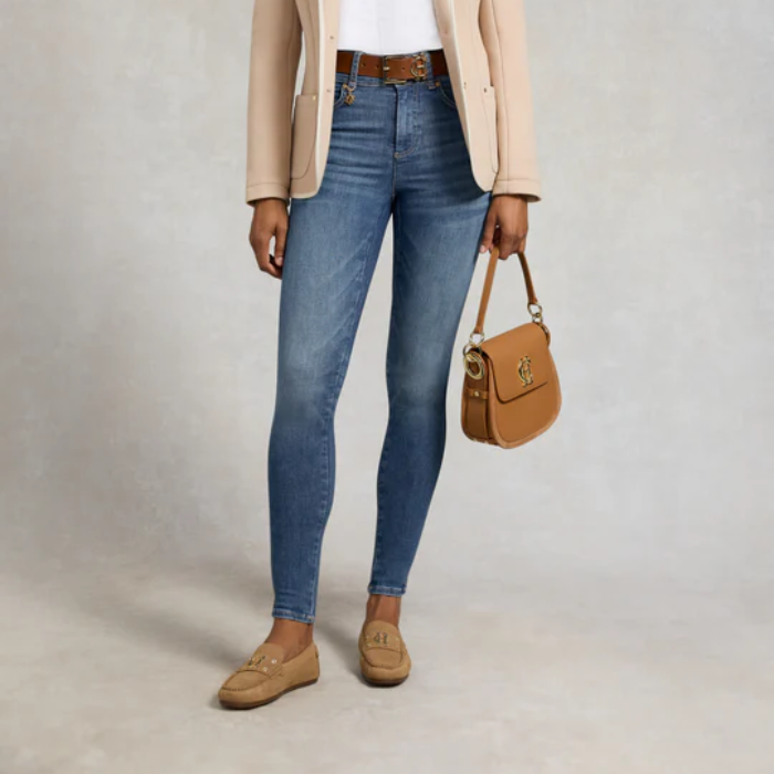 Person wearing blue jeans with a beige jacket and brown shoes, holding a tan handbag.