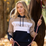 LeMieux AW26 Kadie Colour Block Hoodie Varient Navy. A woman stands outdoors in front of a horse, wearing a color-blocked hoodie with the brand name 'LEMIEUX' printed across the chest. The hoodie features beige, white, and navy blue sections, and she holds a pinkish-purple braided lead rope.