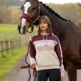 LeMieux AW26 Kadie Colour Block Hoodie Varient Wild Rose. A woman wearing a two-tone burgundy and beige hoodie with 'LEMIEUX' printed on the chest stands next to a brown horse with a white blaze on a path in a field.