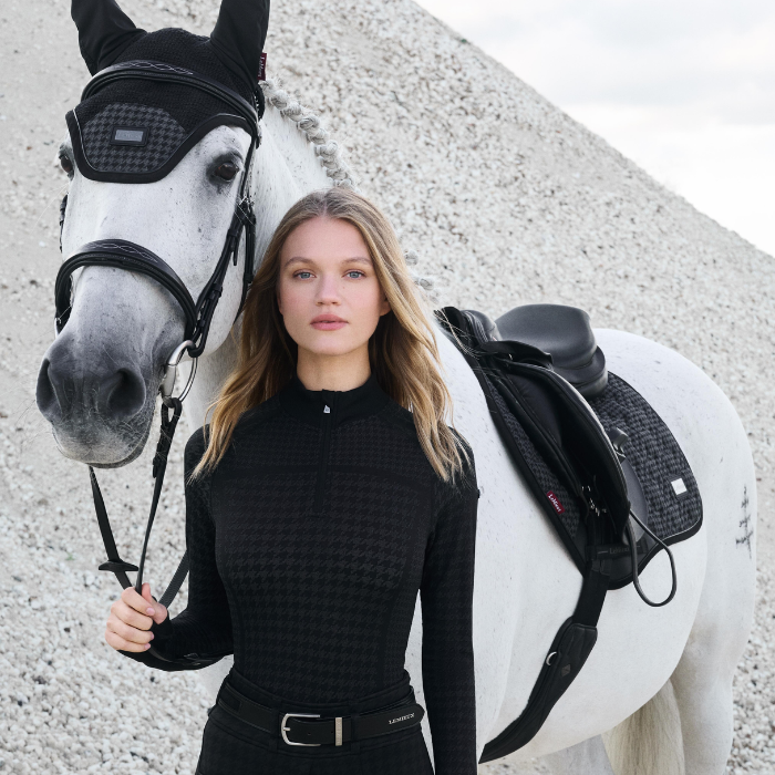 Woman in equestrian attire standing next to a white horse with a black saddle