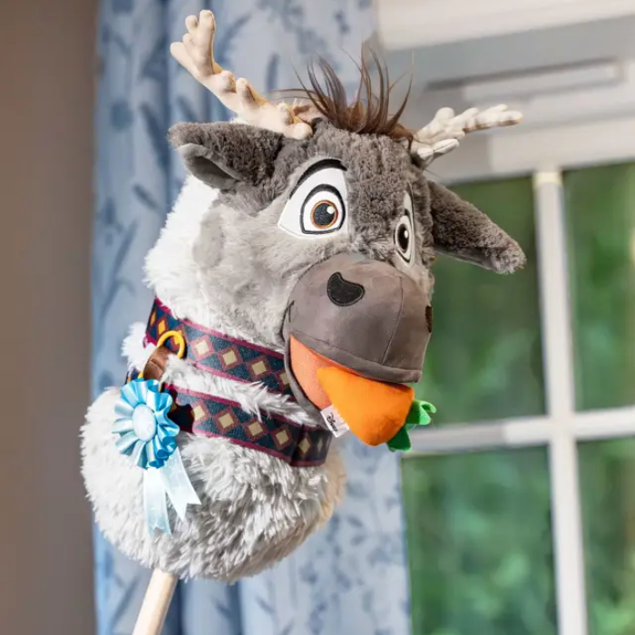 Plush reindeer toy with colorful scarf and carrot in mouth against a blurred indoor background