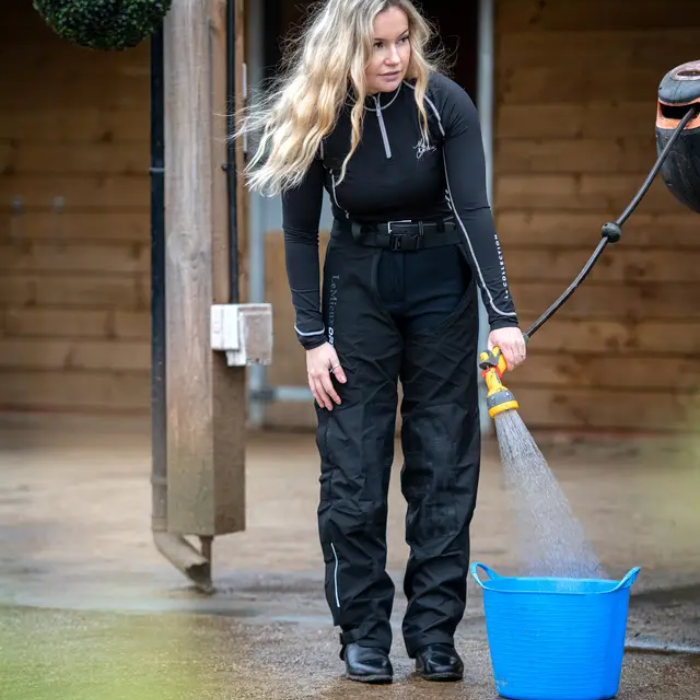 Woman wears a pair of black full chaps and black base layer whilst holding a hose on a yard setting