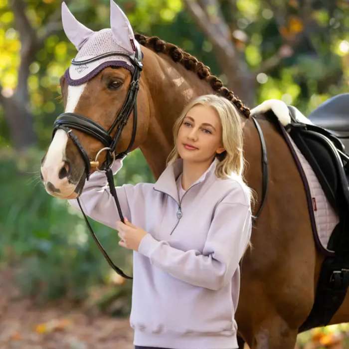 Woman wearing lilac sweatshirt standing next to a horse with a bridle in an outdoor setting