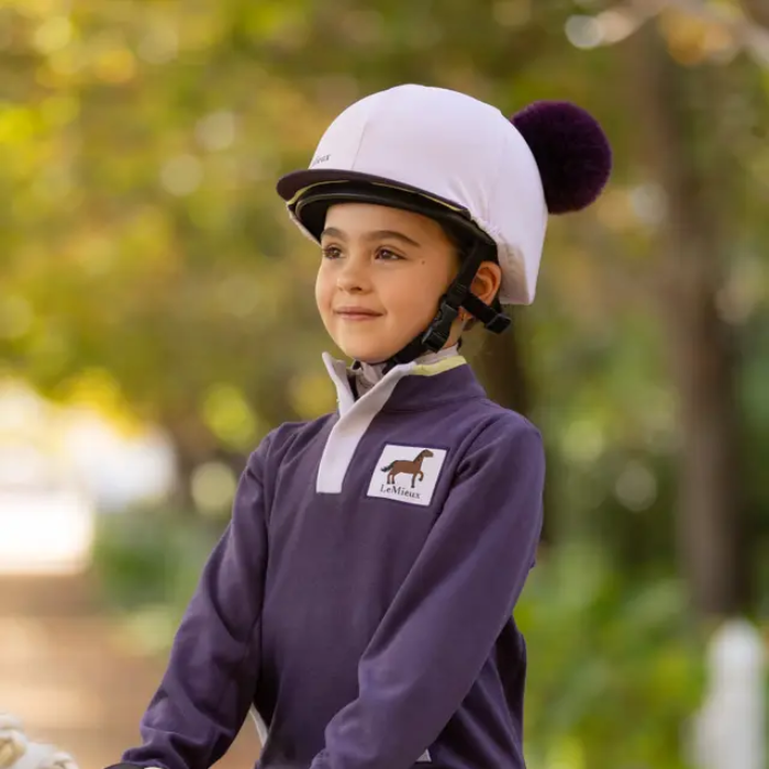 Child in equestrian attire with a helmet, standing outdoors with a blurred natural background