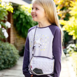 Young girl wearing a purple and grey shirt with a matching crossbody bag outdoors.