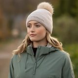 Woman wearing a beige knit hat with a pom-pom and a green jacket outdoors.