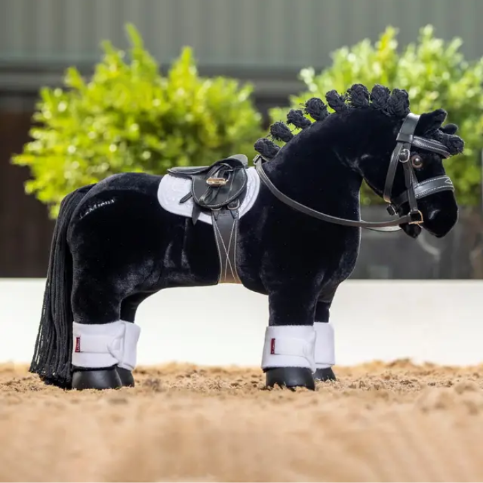 Black plush horse with a white saddle blanket and bridle on a sandy surface.