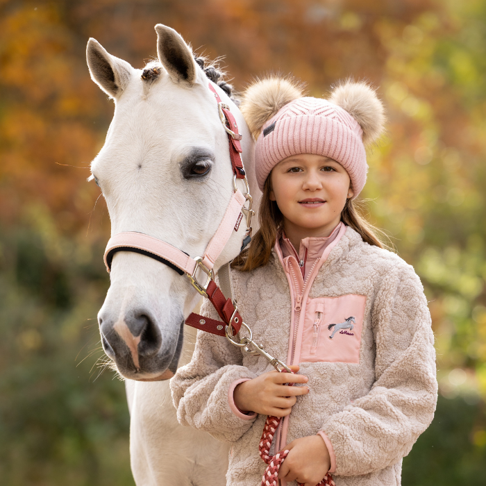LeMieux AW26 Medley Headcollar Variant Colour Spiced Earth/Petal/Aloe. Young person next to a white pony. Pony wearing pink headcollar and person wearing pink wool hat with double pom and pink jacket on faded autumn background.