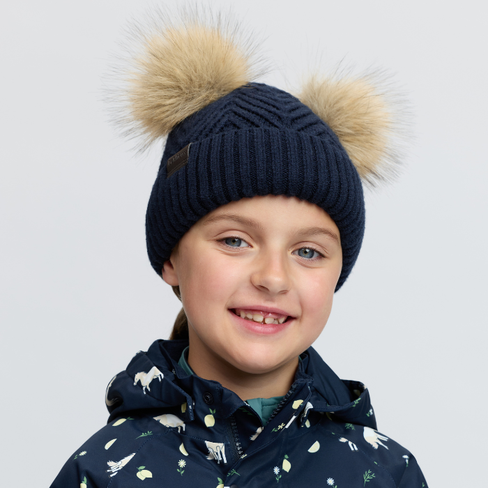 LeMieux AW26 Mini Double Pom Beanie Variant Colour Navy.
A smiling child wearing a navy ribbed knit beanie with two large beige faux-fur pom-poms and a navy patterned winter jacket, photographed against a light gray studio background.