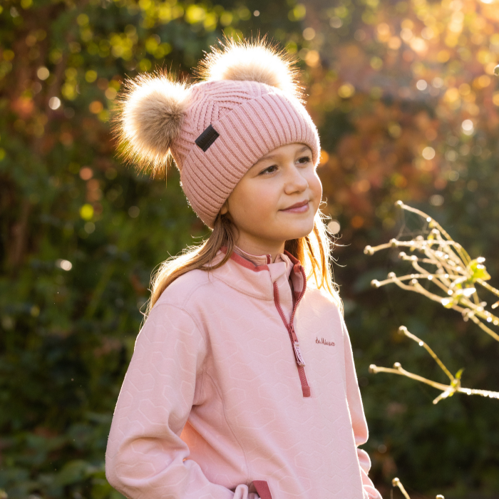 LeMieux AW26 Mini Double Pom Beanie Variant Colour Petal.
A young person wearing a pink ribbed knit beanie with two fluffy pom-poms and a matching pink quarter-zip fleece jacket, photographed outdoors in warm sunlight against a blurred green and gold background.