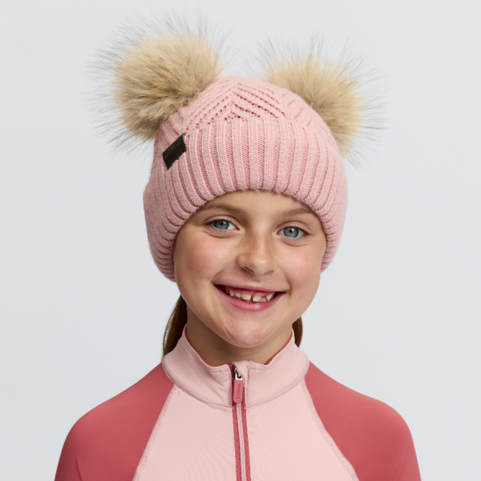 LeMieux AW26 Mini Double Pom Beanie Variant Colour Petal.
A smiling young person wearing a pink ribbed knit beanie with two large beige faux-fur pom-poms and a pink zip-front top, photographed against a light gray studio background.