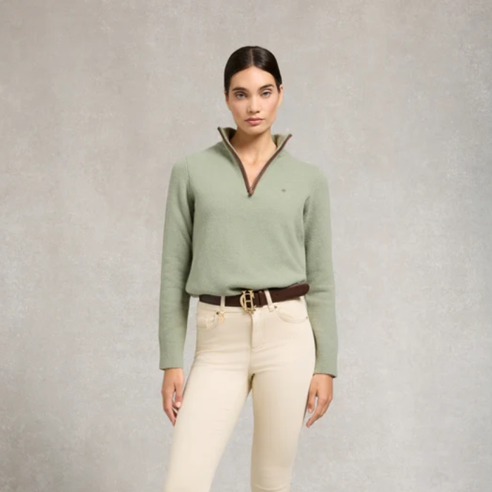 Holland Cooper Country Quarter Zip Knit Variant Colour Sage. A model wearing a sage green sweater, brown belt and cream trousers on grey background.