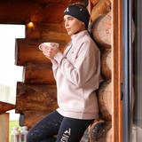 Woman in a beige sweatshirt and black leggings holding a mug, leaning against a wooden wall.