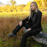 LeMieux AW26 Stormwear Stretch Waterproof Trousers Varient Black. A woman sits on a log in an outdoor grassy field at golden hour, wearing a black technical jacket with 'LEMIEUX' printed vertically on the sleeve and black pants.
