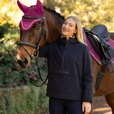 LeMieux AW26 Phoebe Half Zip Waterproof Anorak Varient Navy. A woman stands beside a brown horse outdoors, wearing a dark navy LeMieux equestrian jacket. The horse is outfitted with a magenta and black fly veil and a black saddle with a magenta saddle pad.