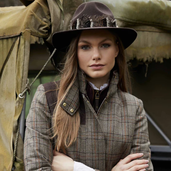 woman in a rural setting wears a brown feathered fedora and tweed coat