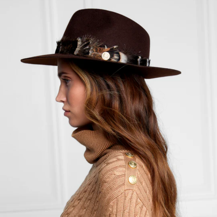 Woman wearing a brown hat with feather accents and a beige cable knit sweater against a white background
