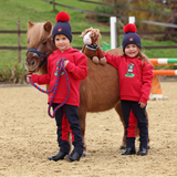 Two children in red sweatshirts and blue hats standing with a brown horse in an outdoor setting.