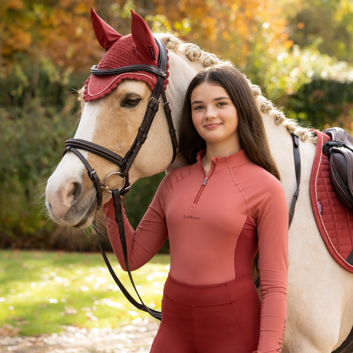 LeMieux AW26 Young Rider Classique Base Layer Variant Colour Spiced Earth.
A young person wearing a rust-colored equestrian quarter-zip shirt and matching breeches stands next to a light-colored horse wearing a matching rust-colored fly veil and bridle. The brand name 'LeMieux' is visible on the shirt.