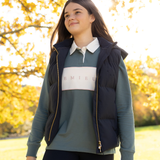 LeMieux AW26 Young Rider Joey Hooded Gilet Varient Navy. A young woman wearing a dark navy puffer vest over a sage green long-sleeve polo shirt with a white chest panel and gold zipper details, standing outdoors in a sunny park with autumn trees.