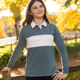 LeMieux AW26 Young Rider Riley Rugby Shirt Varient Aloe. A young woman wearing a slate blue long-sleeve rugby shirt with a white collar and a white chest stripe featuring the brand name 'LEMIEUX' in pink, standing outdoors in a sunny park with autumn trees.