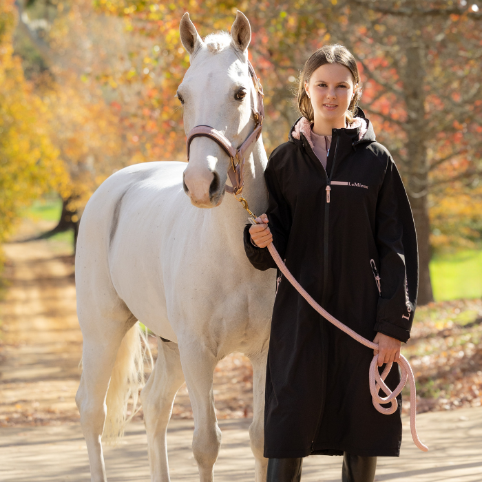 LeMieux AW26 Young Rider HydroShield Robe Varient Black. A woman stands outdoors beside a white horse on a path, wearing a long black coat and holding a pink lead rope. Trees with autumn foliage are blurred in the background.