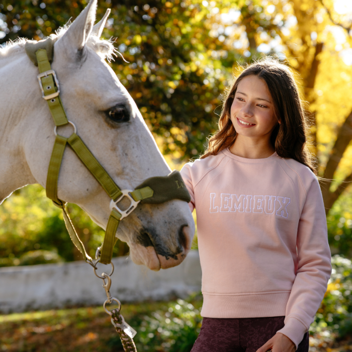 LeMieux AW26 Young Rider Shay Varsity Crew Varient Petal. A smiling young woman stands outdoors beside a white horse wearing an olive green halter. The woman wears a light pink sweatshirt with 'LEMIEUX' printed across the chest in a pale outlined font, paired with dark purple leggings.