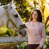 LeMieux AW26 Young Rider Shay Varsity Crew Varient Petal. A smiling young woman stands outdoors beside a white horse wearing an olive green halter. The woman wears a light pink sweatshirt with 'LEMIEUX' printed across the chest in a pale outlined font, paired with dark purple leggings.