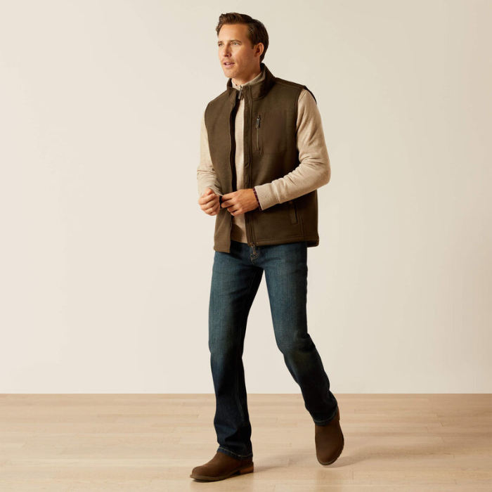 Man wearing a brown gilet, beige shirt, blue jeans, and brown shoes on a plain background