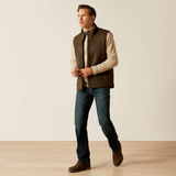 Man wearing a brown gilet, beige shirt, blue jeans, and brown shoes on a plain background