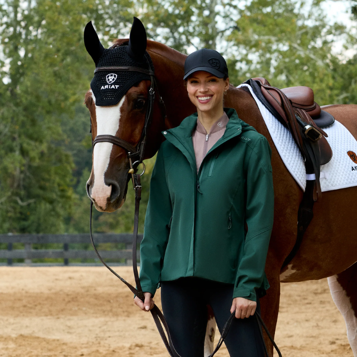 Woman wearing a green coat and blue cap standing next to a horse in an outdoor setting