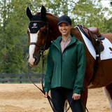 Woman wearing a green coat and blue cap standing next to a horse in an outdoor setting