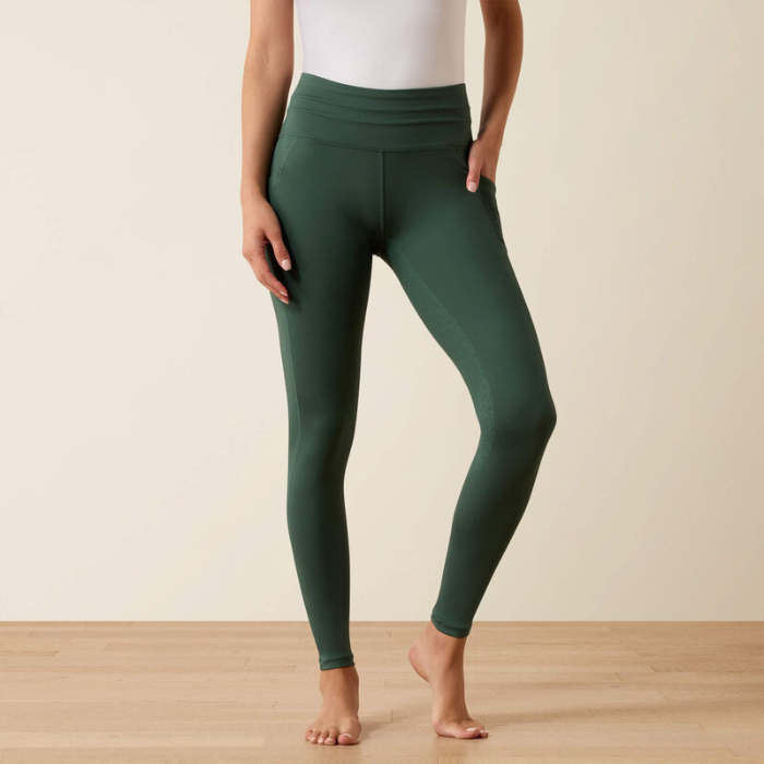 Woman wears a pair of green riding tights and a white top on a beige background
