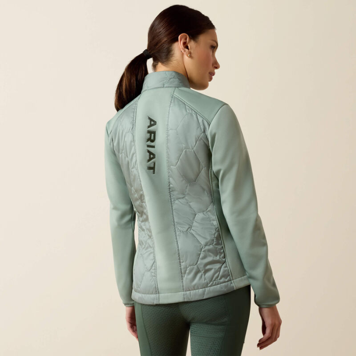 Person wearing a light green quilted jacket with 'Ariat' branding on the back, standing against a beige background.