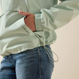 Person wearing a light green jacket with 'Ariat' branding and blue jeans on a plain background