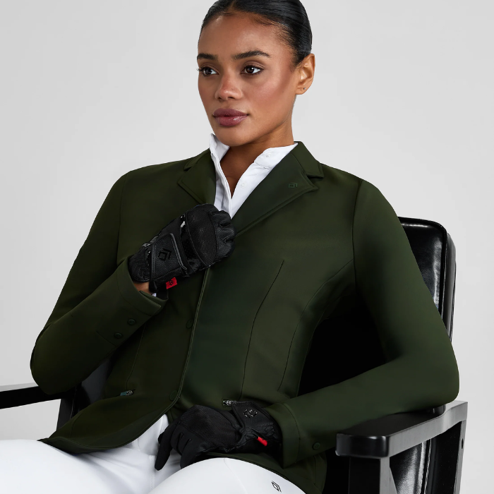 Woman wearing a green equestrian jacket and gloves sitting on a chair.