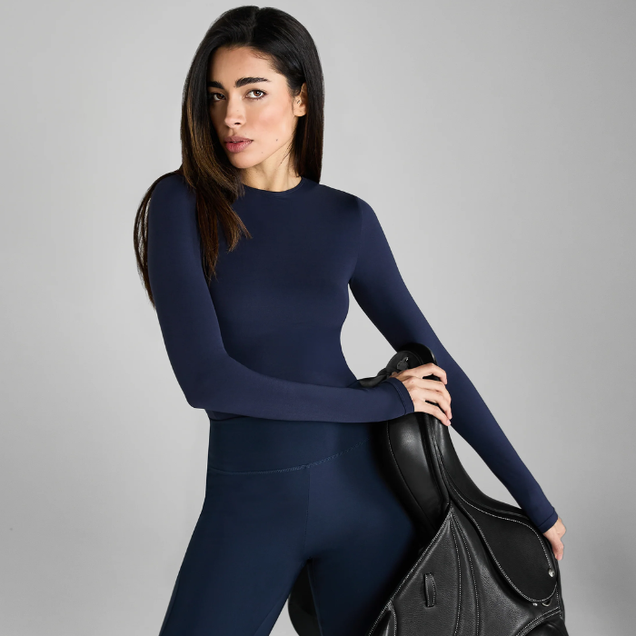 Woman wearing a navy blue outfit holding a black saddle against a grey background