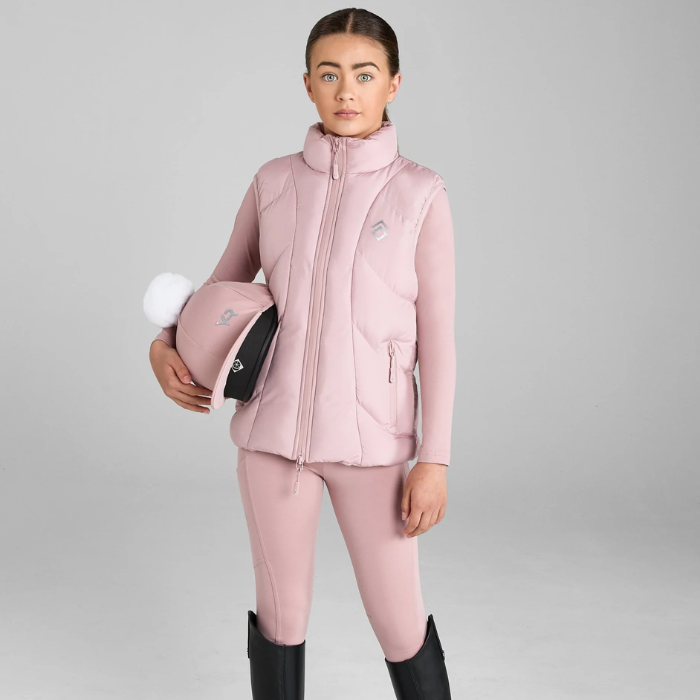 Person wearing a pink equestrian outfit holding a helmet on a grey background. Aztec Diamond Young Rider Mid-Layer Puffer Gilet Pink
