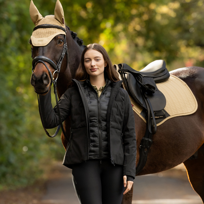 LeMieux AW26 Brooke 3 in 1 Waterproof Hybrid Jacket Varient Black. A woman in a black puffer jacket and black leggings stands beside a brown horse outdoors, wearing tan horse fly blankets on its ears and saddle pad. The background is a soft-focus green outdoor setting.