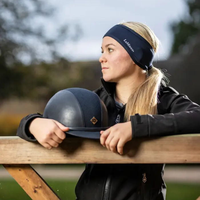 LeMieux AW26 Ear Warmer Headband Variant Colour Indigo. A model with blond hair wearing a blue headband, holding a riding helmet leaning against a gate on faded garden background. 