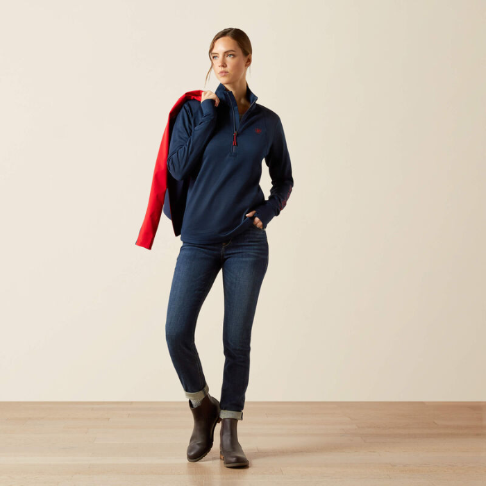 Woman wearing a navy blue jacket with a red hood, blue jeans, and brown boots on a beige background