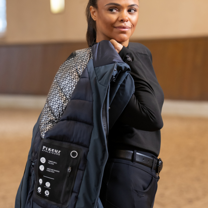 Woman holding a graphite blue jacket over her shoulder with visible branding indoors