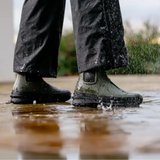 Person wearing green rubber boots walking through a puddle with raindrops falling.