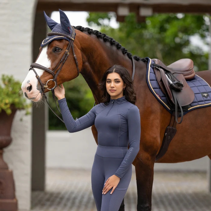 Woman stands next to a horse wearing a dark blue riding outfit in a stable yard area